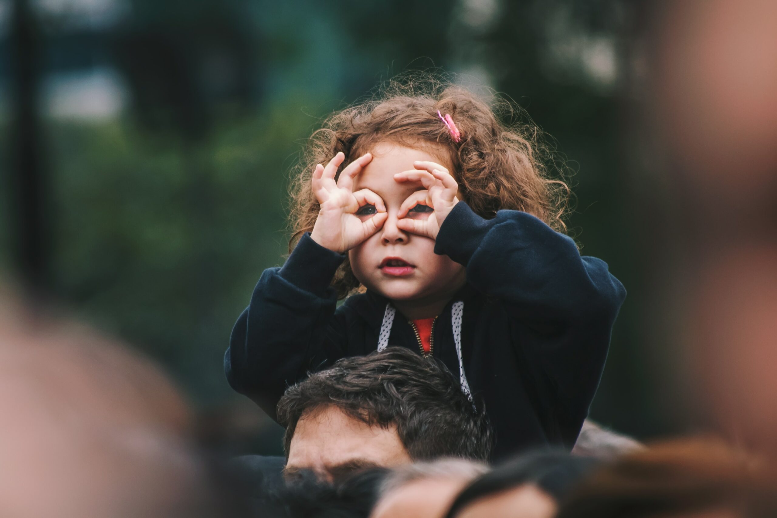 Ein Kind mit schulterlangen, braun-rötlichen Locken sitzt auf den Schultern einer Person. Mit den Händen formt sie zwei Brillengläser und scheint in die Ferne zu blicken.